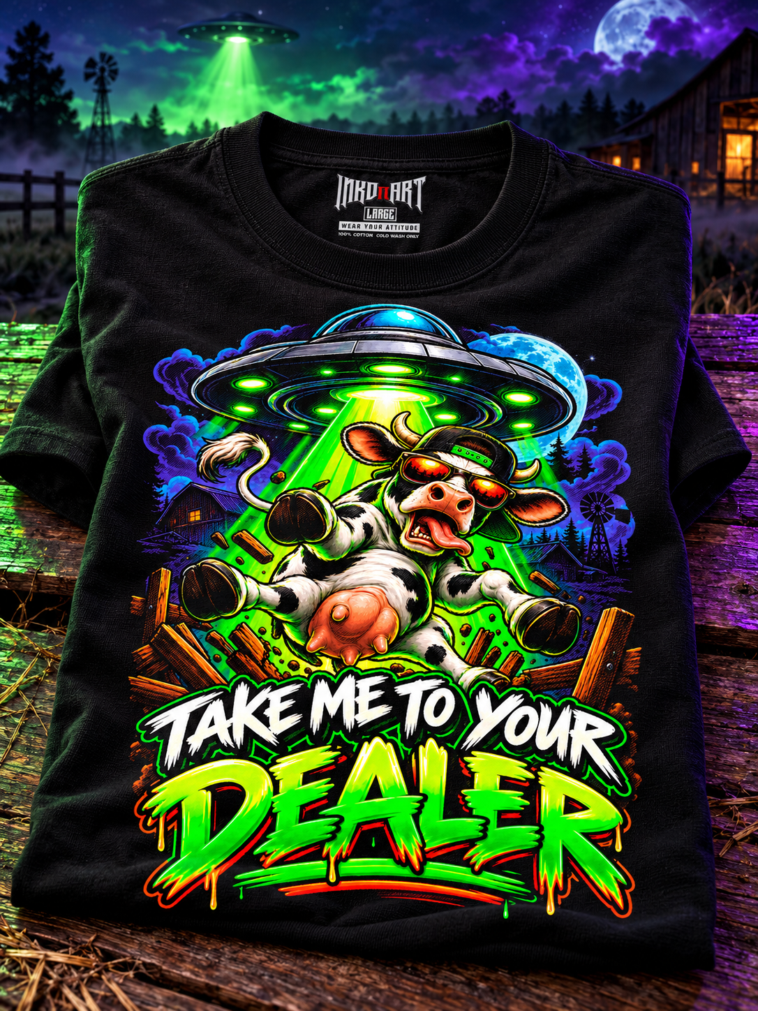 Folded black Take Me to Your Dealer UFO Cow graphic tee on dark barn wood with restrained green and violet lighting, with its printed interior INKDnART neck tag.